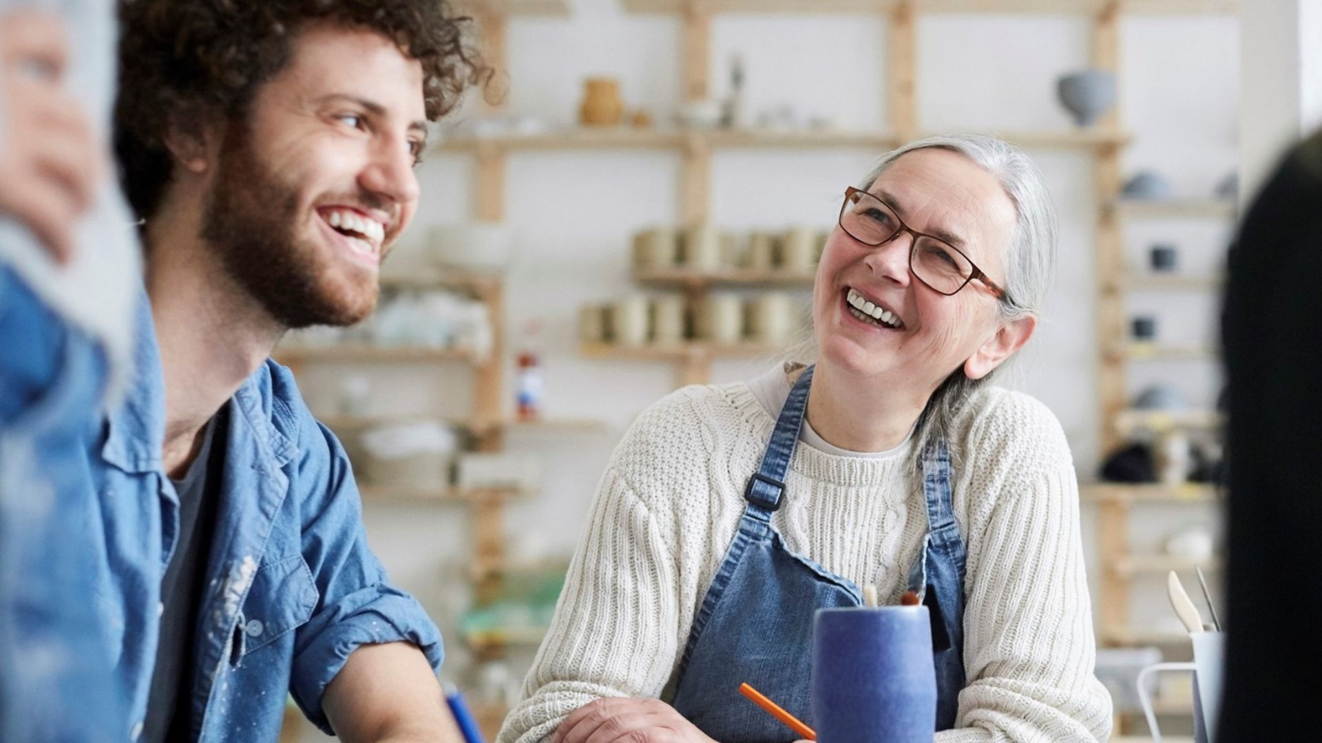 Two people sit at a table, smiling and chatting in a bright room with shelves of pottery and art supplies in the background. One wears glasses and a sweater with an apron, the other has curly hair and a denim shirt.