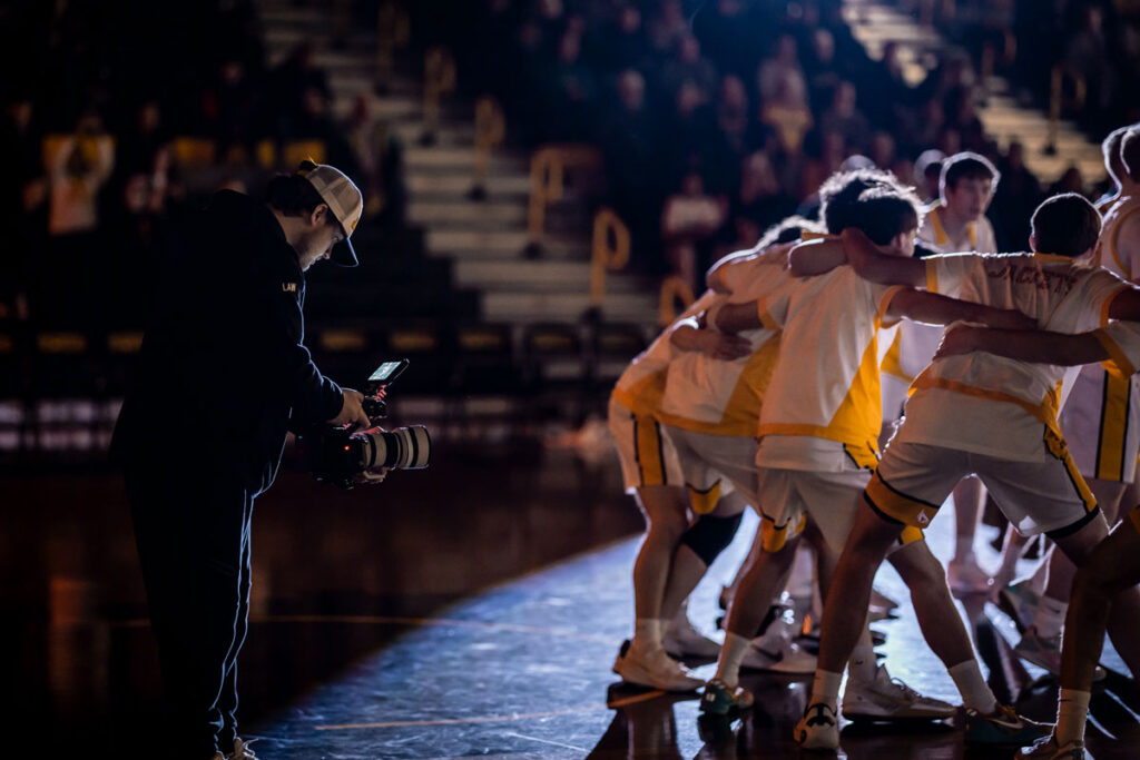 A cameraman, part of a student employment program, films a basketball team in white and yellow uniforms as they huddle on a dimly lit court with spectators watching in the background.