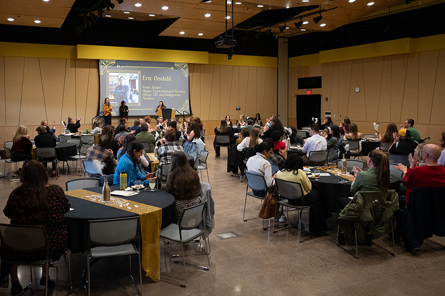 People seated at round tables in a large room, watching a presentation on a projected screen.