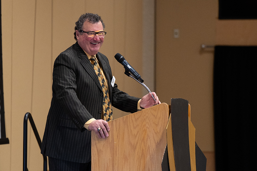 A man in a pinstripe suit and glasses speaks at a wooden podium.