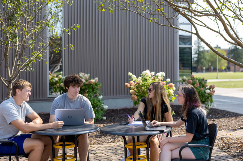 Four students sit outside at two round tables, working on laptops and taking notes. Surrounded by plants and trees near a modern UW-Superior building, they enjoy a sunny day—perhaps discussing graduate programs or online degrees.