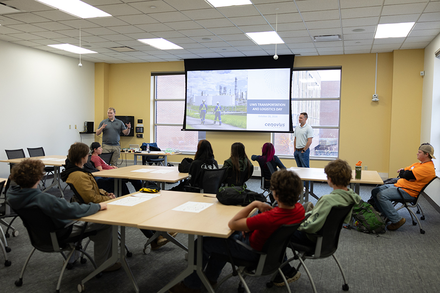 A speaker presents to a group of students in a classroom with a large projected slide.
