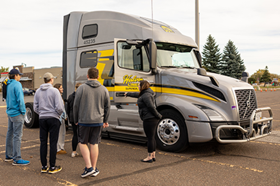 Six people stand near a large silver semi-truck with yellow and black stripes in a parking lot, looking at and discussing the vehicle on a clear day with trees and buildings in the background.