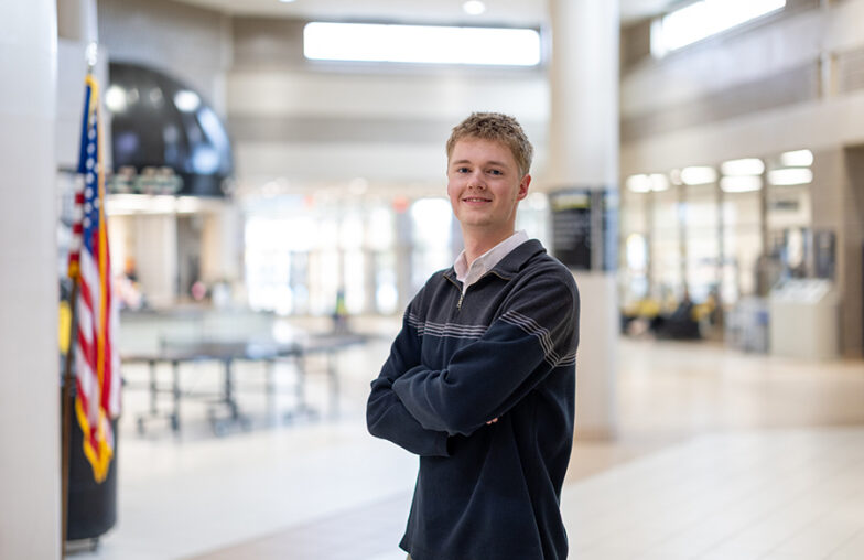A young man stands with arms crossed in a bright, modern building near an American flag.