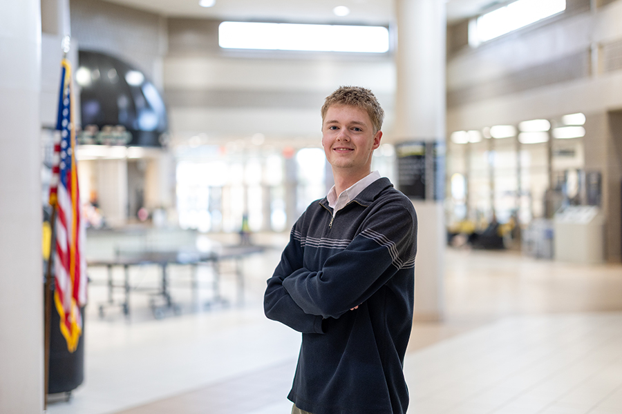 A young man stands with arms crossed in a bright, modern building near an American flag.