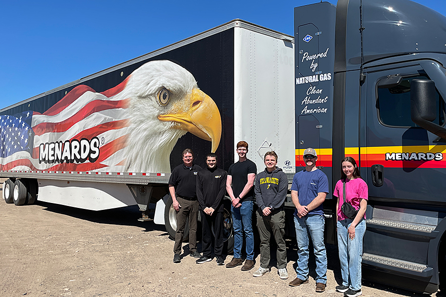 Six people stand in front of a Menards truck featuring an eagle and an American flag design.