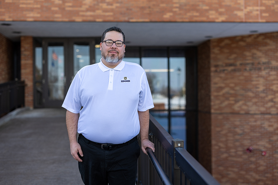 A man in a white polo shirt stands outside a brick building, holding a railing.