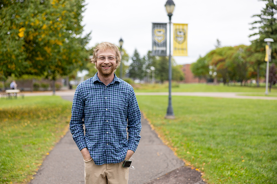 A smiling man with glasses and a blue plaid shirt stands on a campus path.