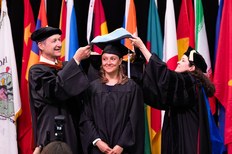 A graduate smiles as two faculty members place a graduation hood on her during the graduation ceremony. They stand before a display of international flags, all dressed in academic gowns and caps.