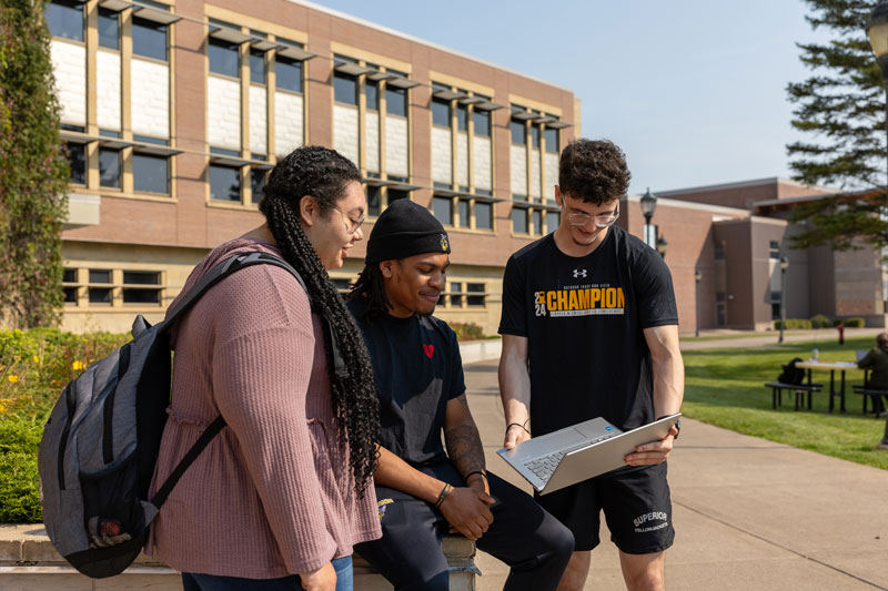 Three college students stand together outside on campus, looking at a laptop and smiling. A brick academic building and trees are visible in the background on a sunny day.