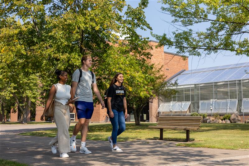 Three students walk together outside on a sunny day, smiling, with trees, a bench, and a brick building with large windows in the background.