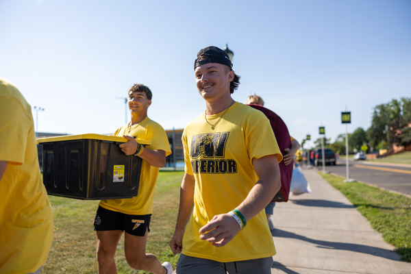 Two young men wearing yellow UW-Superior shirts walk outside on a sunny day, moving to campus. One carries a large black bin as they appear cheerful, accompanied by others in the background.