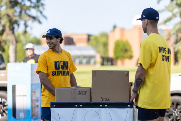 Two young men wearing yellow shirts, one with UW Superior and the other with Move-in Crew, stand outside with cardboard boxes on a sunny day, helping students moving to campus during a move-in event.
