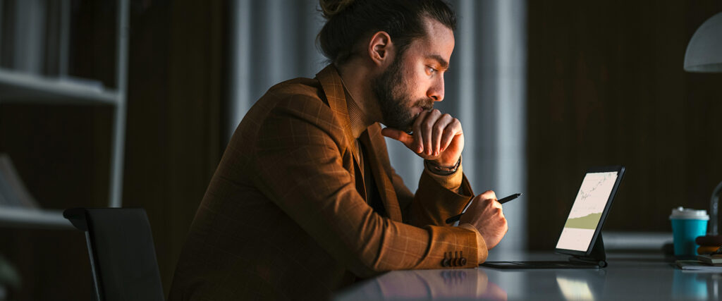 A man in a brown plaid jacket sits at a desk, looking thoughtfully at a tablet with a stylus in hand learning about the Finance degree at UW-Superior.