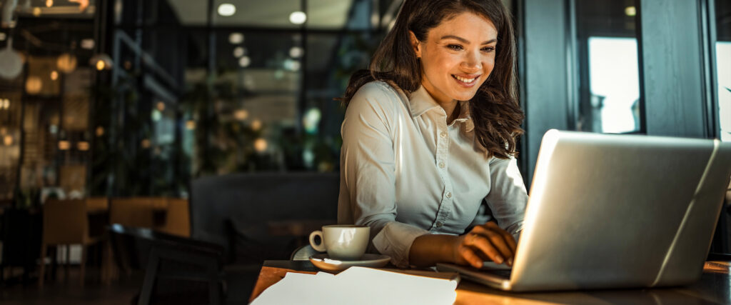 Smiling woman in a white shirt sits at a table in a café, working on a laptop,.