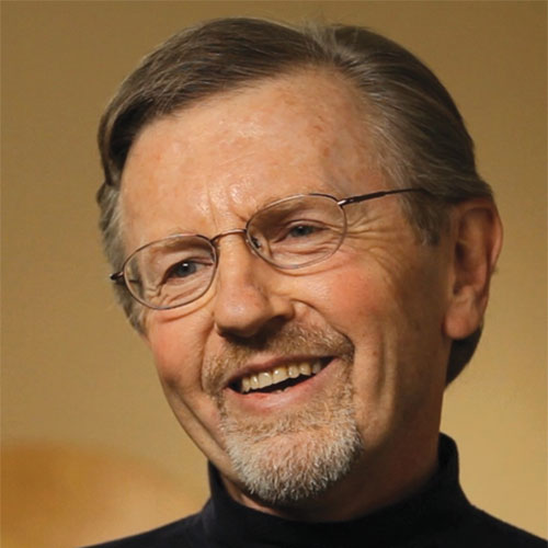 A smiling older man with short gray hair, glasses, and a goatee, wearing a dark turtleneck, appears to be speaking or laughing about past events and activities against a softly lit, neutral background.