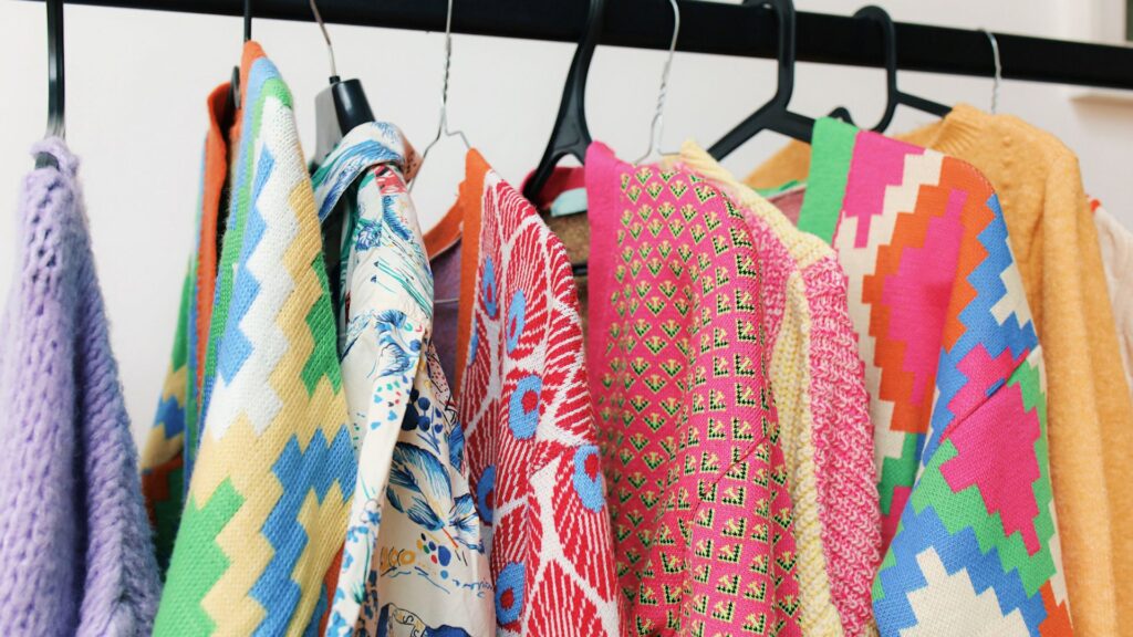 A row of colorful sweaters and shirts with various bold patterns and textures hanging on black hangers on a clothing rack.