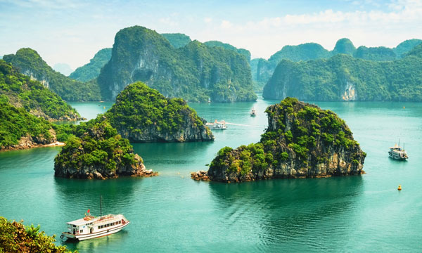 A scenic view of Ha Long Bay in Vietnam, featuring emerald green water, lush, rocky islands, and several boats cruising among the islands under a clear sky.