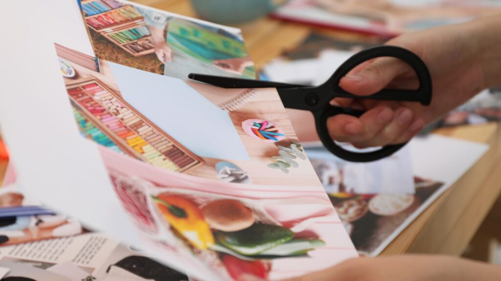 A person uses black scissors to cut out colorful images from a magazine page, possibly for a collage or craft project, on a wooden table scattered with more clippings and materials.