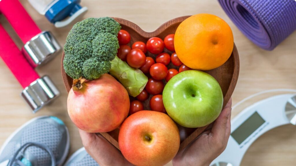 A person holds a heart-shaped bowl filled with broccoli, cherry tomatoes, an orange, an apple, and a pomegranate. Around it are sneakers, a yoga mat, dumbbells, and a bathroom scale, symbolizing healthy living.