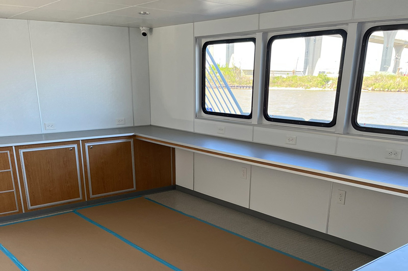 The interior of a boat or ship cabin is about spacious comfort, featuring large windows, built-in cabinets, and a long countertop. The floor is covered with protective cardboard sheets. Water and greenery are visible through the windows.