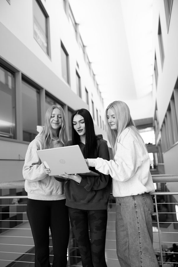 Three young women stand indoors, smiling and enjoying open communication while viewing a laptop.