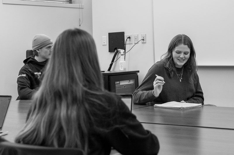 Three students studying legal studies sit at a table, one writing in a notebook and smiling.
