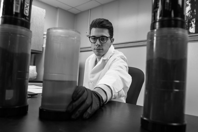 Person in lab coat and gloves handling a large container in a chemical engineering laboratory.