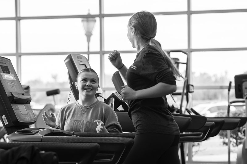 Two women discuss health promotion and wellness while using treadmills in a bright gym.