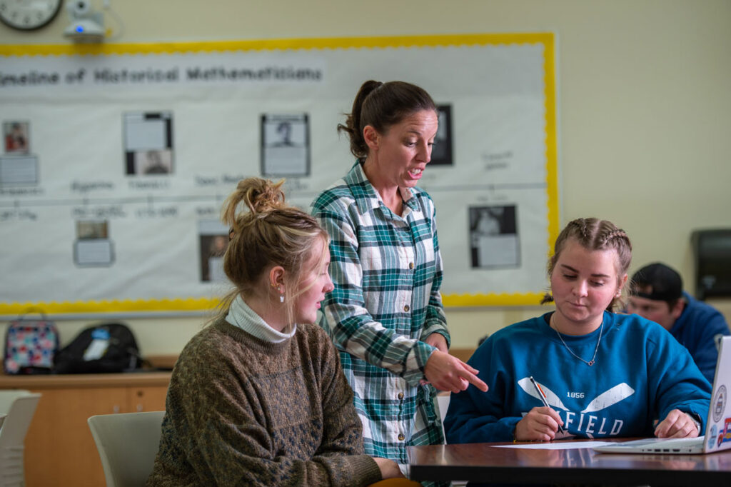 A teacher stands and points at a notebook while discussing english education with two students seated at a desk in a classroom. One student writes while the other listens. A bulletin board and timeline are visible in the background.