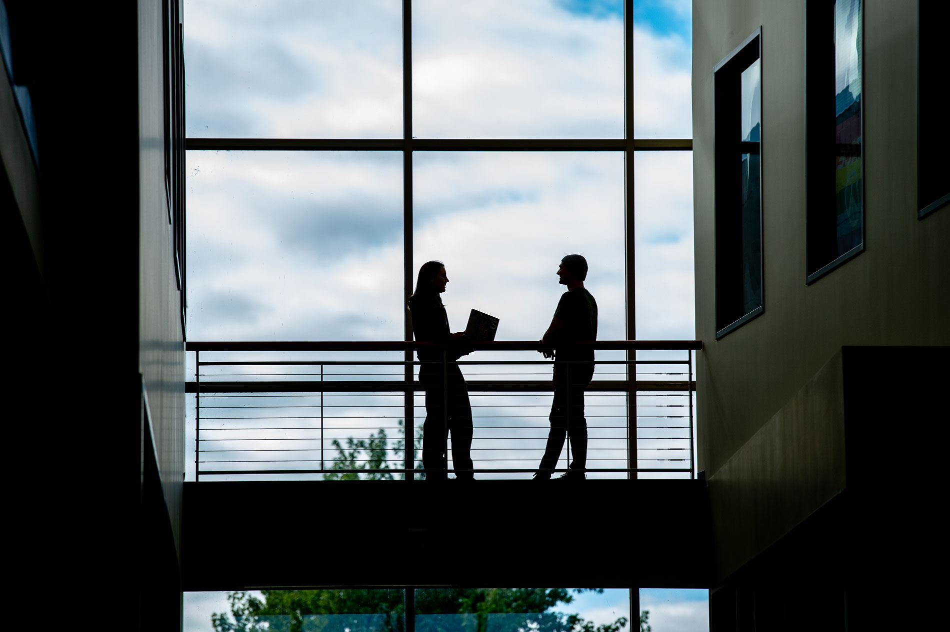 Two people in discussion on an indoor walkway with large windows and a cloudy sky.