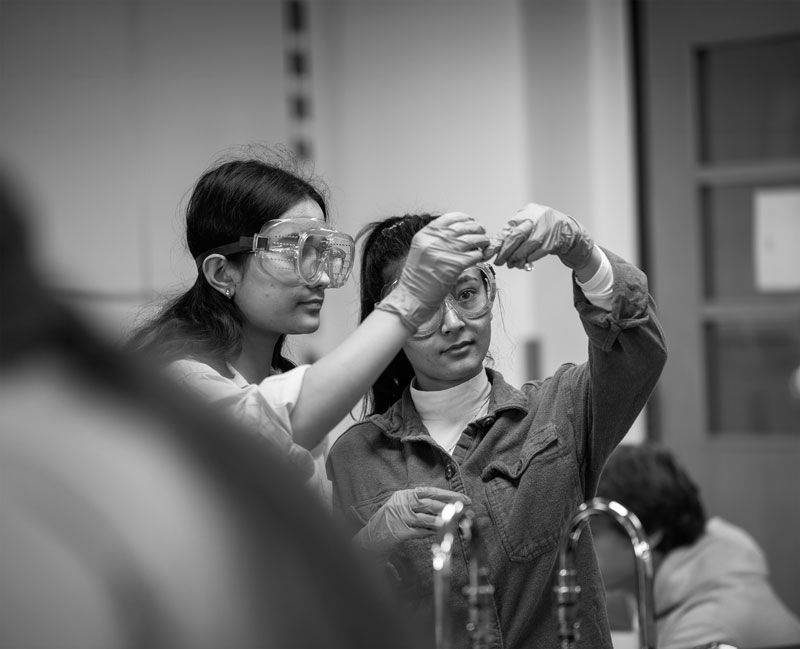 Two people in goggles and gloves examine a small object in a pre-medicine laboratory setting.
