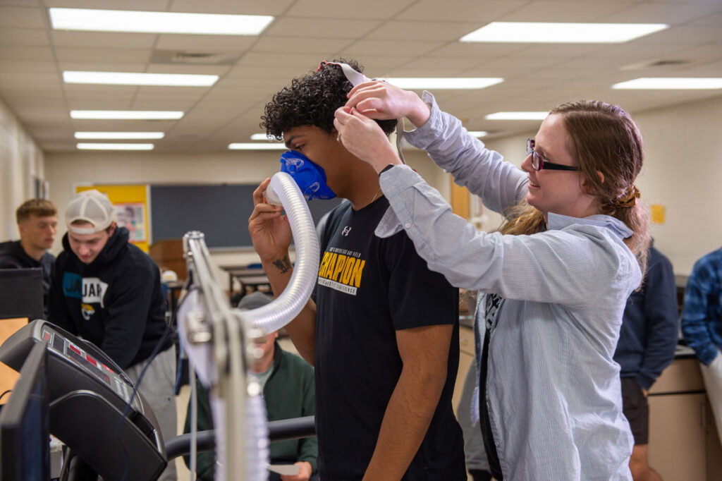 A woman adjusts a head strap for a man wearing a blue oxygen mask connected to a machine, possibly for a fitness or respiratory test in a classroom—perhaps as part of health minor coursework—with other students in the background.