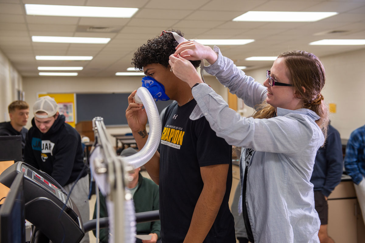 A woman adjusts a head strap for a man wearing a blue oxygen mask connected to a machine, possibly for a fitness or respiratory test in a classroom—perhaps as part of health minor coursework—with other students in the background.