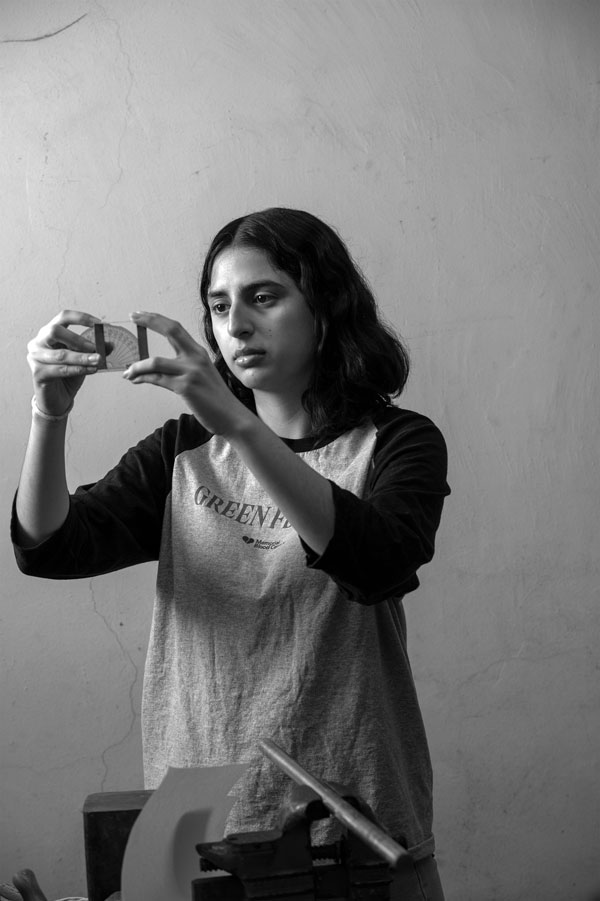 A student examining a small object with both hands, reflecting on art and design, against a plain wall.