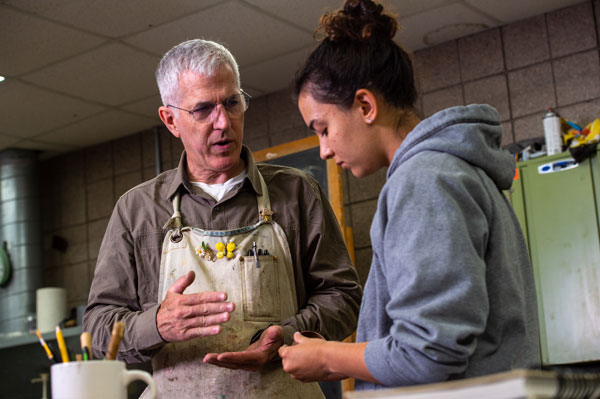 An older man in an apron shares fast facts with a young woman in a gray hoodie as they stand together in a workshop or classroom, surrounded by art supplies and tools in the background.