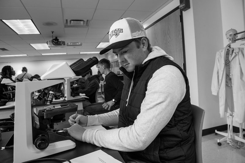 Student uses a microscope in a biology classroom, writing notes; others work in the background.