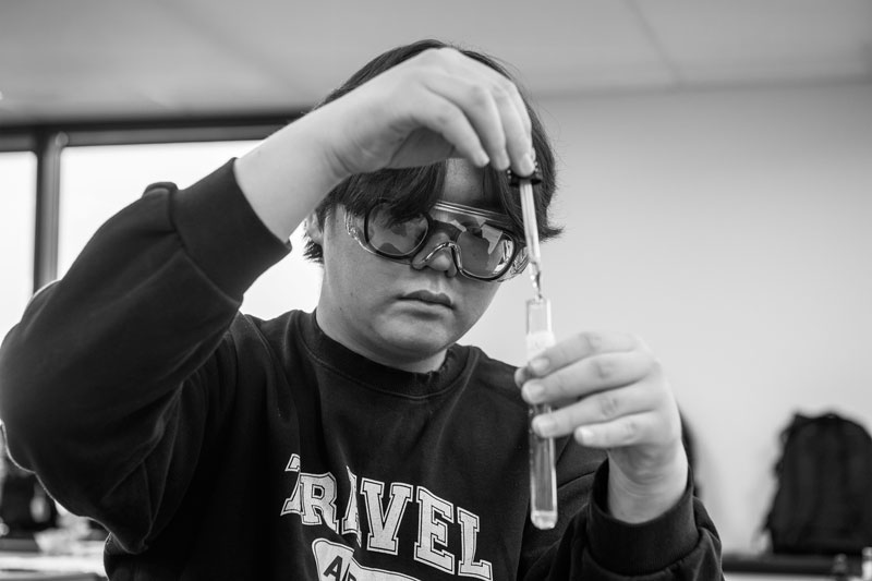 Person wearing goggles performs chemistry, adding liquid to a test tube with a dropper in a lab.