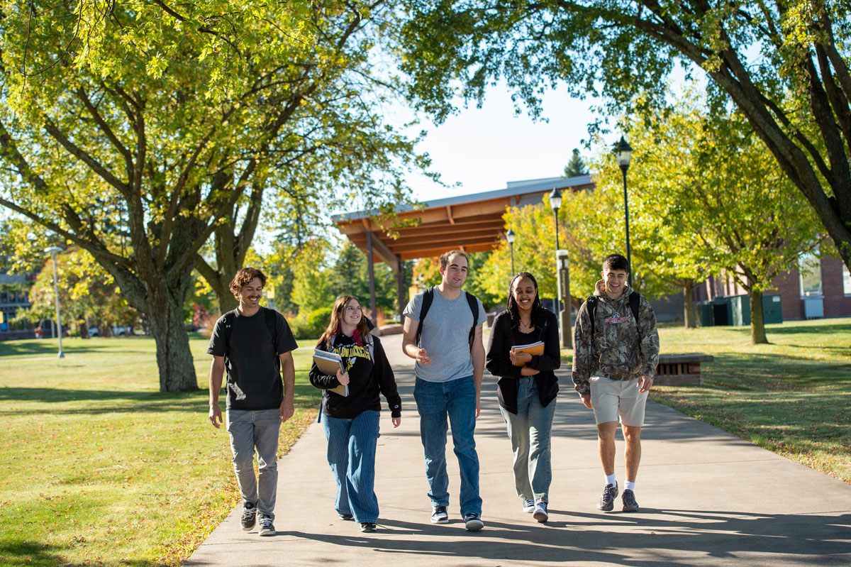 Five students walk together on a sunny University of Wisconsin-Superior campus pathway, surrounded by green trees and grass, carrying backpacks and books, smiling and talking about their program.