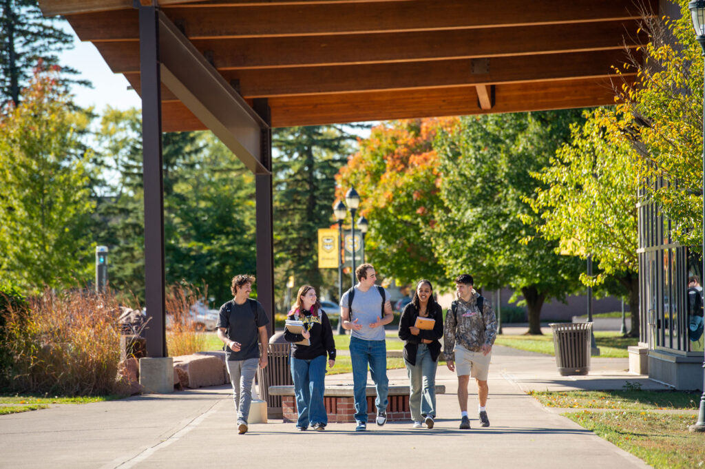 Five students walk together under a large outdoor canopy on a sunny day, surrounded by trees with green and autumn-colored leaves. Smiling and talking about academics, they carry books and backpacks as they enjoy the campus atmosphere.