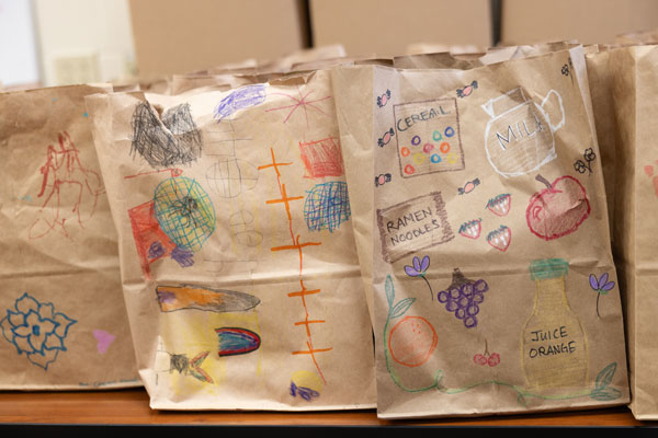 Brown paper bags with food pantry labels and colorful drawings of food items on a wooden surface.