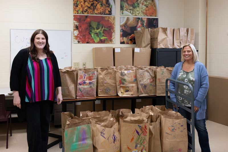 Two women stand by a table with decorated bags and boxes at a food pantry, food photos behind.