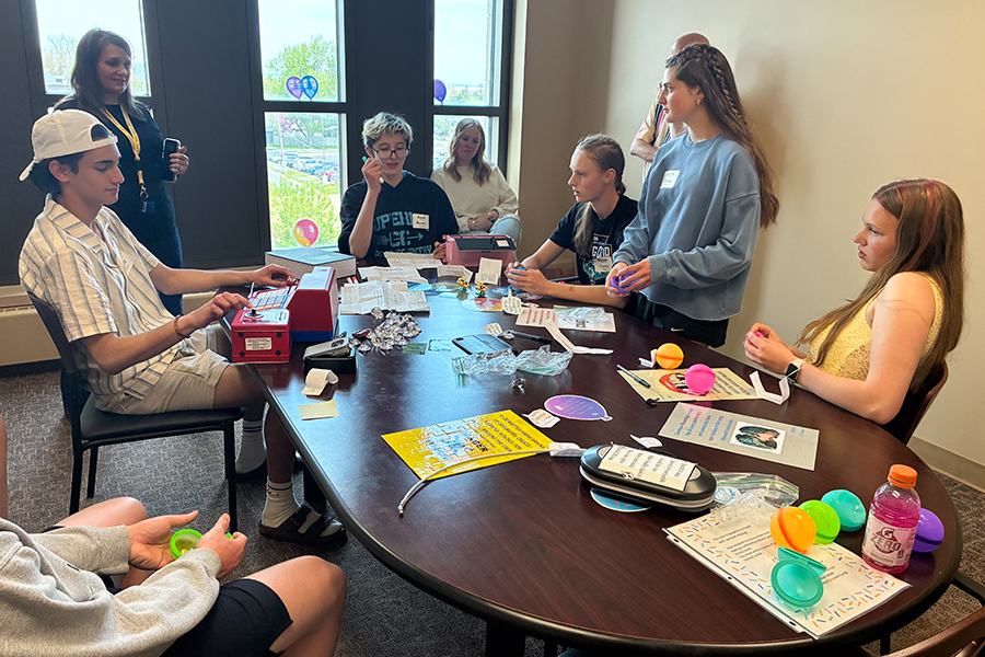 Seven people sit and stand around a table with papers, drinks, and colorful plastic eggs.