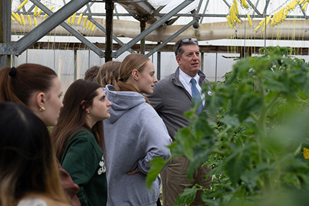 A group of people listen to a man speak inside a greenhouse with green plants visible.