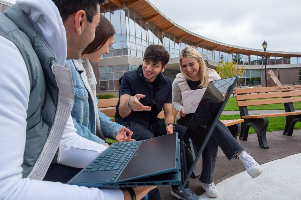 Four students sit on benches outdoors, smiling and talking while using laptops and papers, possibly discussing admissions requirements, with a modern building and green lawn in the background.
