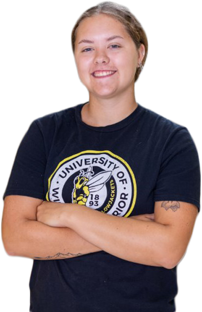 A student with short hair smiles with arms crossed, wearing a black T-shirt featuring a yellow bee and the words University of Wisconsin-Superior. Tattoos are visible on both forearms.