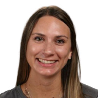 A woman with long straight brown hair and a wide smile, wearing a grey top, pictured against a plain white background.