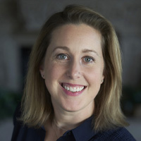A woman with shoulder-length blonde hair and blue eyes smiles at the camera. She is wearing a navy blue top and is in a softly blurred indoor setting.