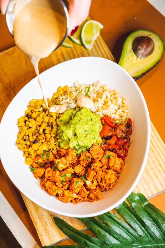 A dining bowl filled with brown rice, roasted corn, diced tomatoes, seasoned plant-based protein, guacamole, and sauce being poured on top, served with half an avocado, a lime wedge, and a green leaf nearby.