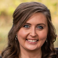 A woman with wavy brown hair and blue eyes smiles at the camera. She is wearing silver earrings and standing outdoors with a blurred natural background.